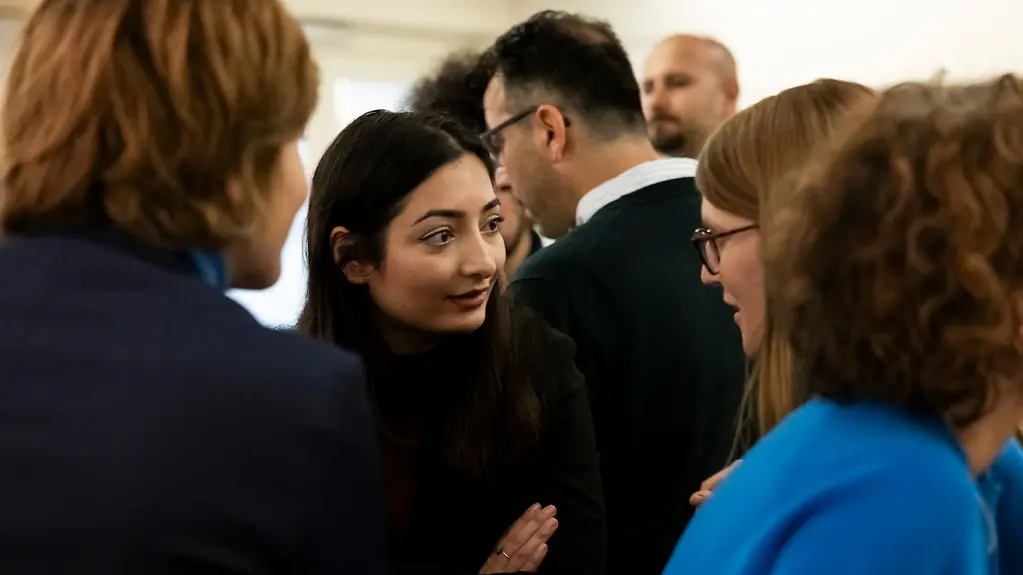 Die Staatsministerin steht in einer Gruppe Menschen und hört sehr neugierig zu.